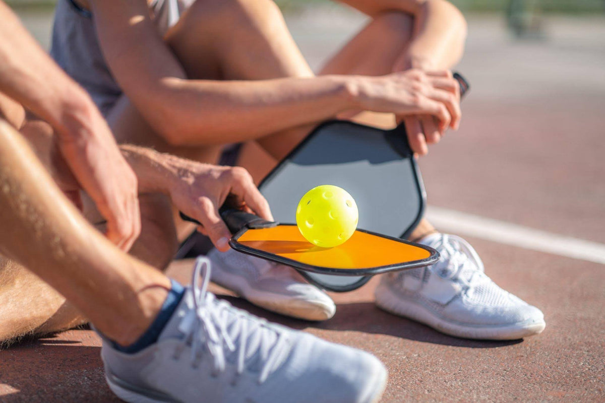 A close-up lifestyle shot of a person sitting on a running track, holding a paddle with a yellow tennis ball on it. Another person's legs and hands are visible in the foreground, and both are wearing white athletic shoes.